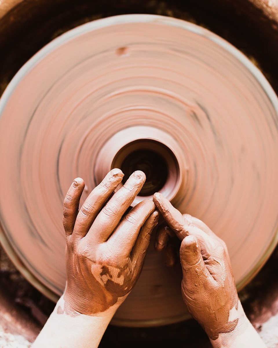 Clay-covered hands shaping a pot on a spinning wheel, seen from above.